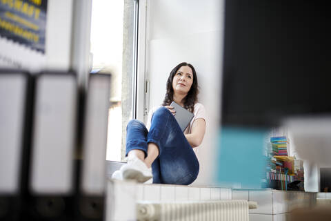 Reife Geschäftsfrau mit digitalem Tablet, während sie auf der Fensterbank im Büro sitzt, lizenzfreies Stockfoto