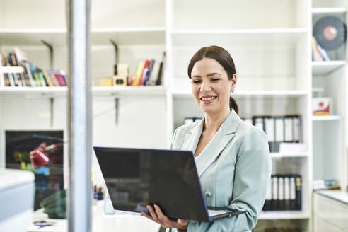 Lächelnde Geschäftsfrau in formeller Kleidung mit Laptop im Büro - MMIF00226