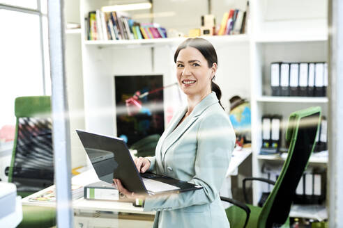 Smiling businesswoman with laptop seen through glass at office - MMIF00228