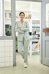 Geschäftsfrau in voller Länge, stehend mit verschränkten Armen an der Tür eines Büros - MMIF00234