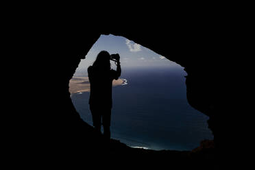 Silhouette eines Mannes, der ein Foto von einer Höhle in den Famara-Felsen aus macht. - CAVF80721