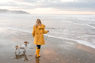 Tween in gelbem Mantel führt Hund am Strand in Neuseeland aus - CAVF80734