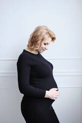 Schwangere Frau im schwarzen Kleid lächelnd berührt Bauch - CAVF80764