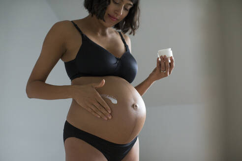 Schwangere Frau in BH und Höschen trägt Feuchtigkeitscreme auf den Bauch auf - HOXF06226