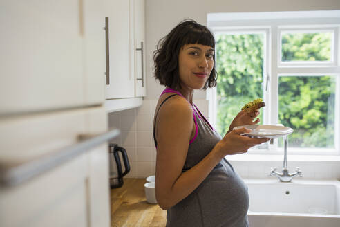 Porträt selbstbewusste schwangere Frau isst Avocado-Toast in der Küche - HOXF06231