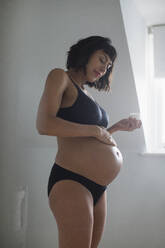 Schwangere Frau in BH und Höschen trägt Feuchtigkeitscreme auf den Bauch auf - HOXF06232