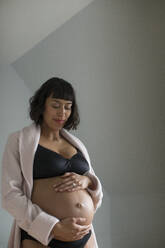 Happy pregnant woman kneeling on the floor in a studio, smiling at her  beautiful baby bump. Young mom-to-be wearing lingerie, cherishing the life  growing inside her with maternal love. stock photo