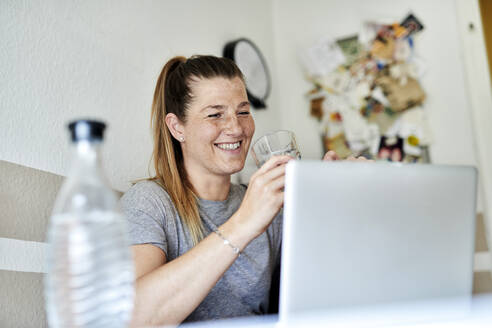 Lächelnde Frau, die Wasser trinkt und einen Laptop im Wohnzimmer benutzt - MMIF00237