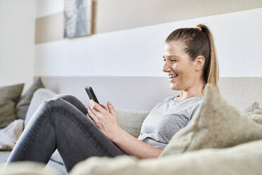 Smiling woman using smart phone while sitting on sofa at home - MMIF00241