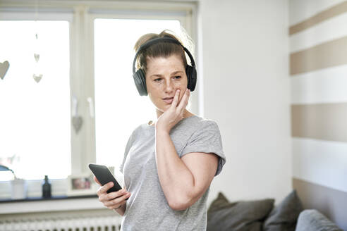 Woman listening through headphones while using smart phone at home - MMIF00243