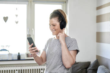 Mittlere erwachsene Frau hört Musik über Kopfhörer, während sie zu Hause ein Mobiltelefon benutzt - MMIF00245