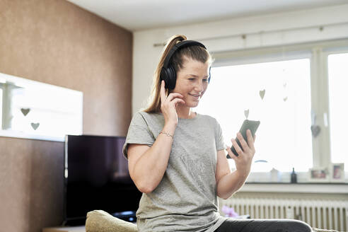 Frau mit Kopfhörern, die zu Hause über ihr Mobiltelefon Musik hört - MMIF00247