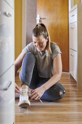 Lächelnde Frau bindet Schnürsenkel, während sie zu Hause auf dem Hartholzboden sitzt - MMIF00249
