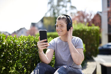 Lächelnde Frau hört Musik über Kopfhörer am Smartphone bei Pflanzen - MMIF00250