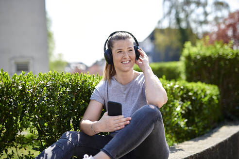 Lächelnde Frau, die wegschaut, während sie Musik über Kopfhörer auf dem Smartphone hört, bei Pflanzen - MMIF00251