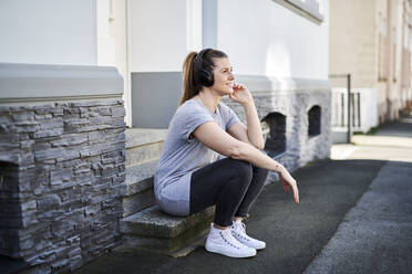 Woman listening music on wireless headphones while sitting on steps - MMIF00252