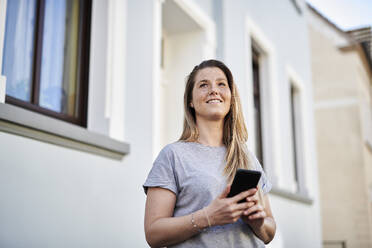 Woman looking away while holding mobile phone against house - MMIF00253
