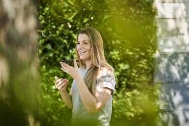 Lächelnde Frau spricht über Lautsprecher mit ihrem Smartphone, während sie inmitten von Pflanzen im Hinterhof steht - MMIF00254