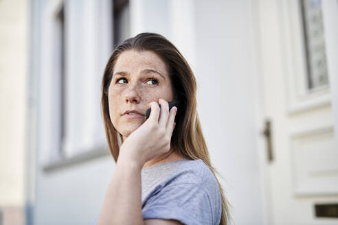 Woman looking away while talking on smart phone against house - MMIF00255