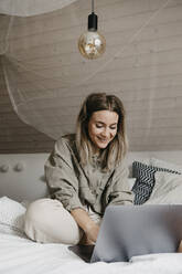 Lächelnde Frau, die einen Laptop benutzt, während sie zu Hause auf dem Bett sitzt - LHPF01273
