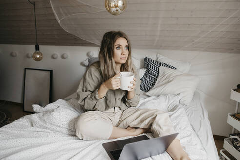 Nachdenkliche Frau mit Laptop trinkt Kaffee, während sie zu Hause auf dem Bett sitzt - LHPF01276