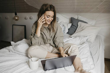 Lächelnde Frau, die mit ihrem Smartphone spricht, während sie zu Hause im Schlafzimmer am Laptop arbeitet - LHPF01278