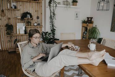 Junge Frau, die einen Laptop benutzt, während sie mit hochgelegten Füßen in einem Haus am Tisch sitzt - LHPF01281