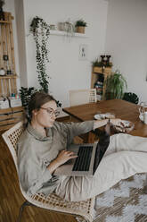 Woman using laptop while sitting at table with feet up in home office - LHPF01282