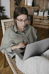 Ernste Frau, die einen Laptop benutzt, während sie auf einem Stuhl zu Hause sitzt - LHPF01283