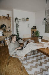 Müde Frau mit hochgelegten Füßen, die einen Laptop benutzt, während sie am Tisch im Wohnzimmer sitzt - LHPF01284