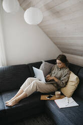 Frau, die einen Laptop benutzt, während sie mit einem Getränk und einem Buch zu Hause auf dem Sofa sitzt - LHPF01285
