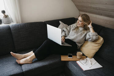 Smiling woman using laptop on sofa while freelancing at home - LHPF01287