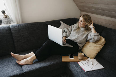 Lächelnde Frau, die einen Laptop auf dem Sofa benutzt, während sie zu Hause freiberuflich arbeitet - LHPF01287