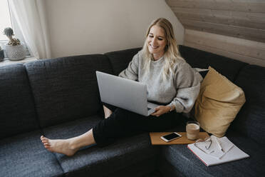 Smiling freelancer using laptop while sitting on sofa with drink and document at home - LHPF01289