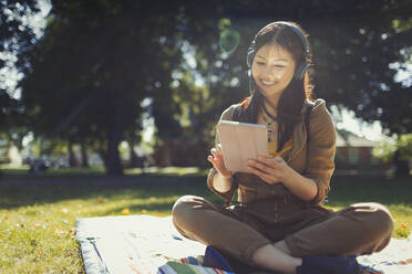 Lächelnde junge Frau, die ein digitales Tablet benutzt und mit Kopfhörern im sonnigen Sommerpark Musik hört - CAIF26292