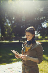Lächelnde junge Frau mit Kopfhörern und digitalem Tablet in einem sonnigen Sommerpark - CAIF26294
