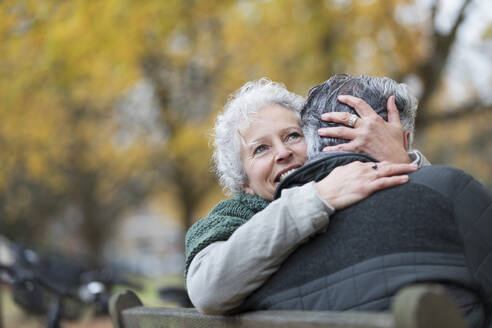 Lächelndes, zärtliches älteres Paar, das sich auf einer Bank im Herbstpark umarmt - CAIF26377
