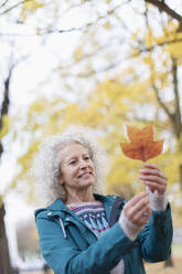 Neugierige ältere Frau hält orangefarbenes Herbstblatt im Park - CAIF26385