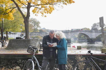 Senior couple with bicycles traveling, looking at guidebook along autumn river - CAIF26388