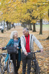 Senior couple walking bicycles among trees and leaves in autumn park - CAIF26394