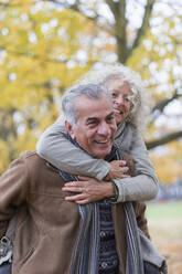 Verspieltes, lächelndes älteres Paar nimmt im Herbst im Park Huckepack - CAIF26395
