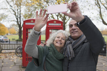 Lächelndes älteres Paar, das ein Selfie im Herbstpark vor einer roten Telefonzelle macht - CAIF26405