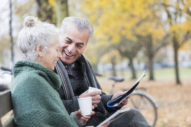 Lächelndes älteres Paar liest Zeitung und trinkt Kaffee auf einer Bank im Herbstpark - CAIF26418