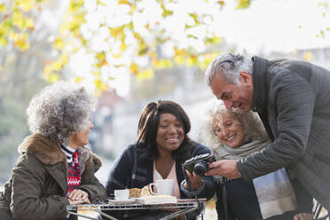 Active senior friends using digital camera at autumn sidewalk cafe - CAIF26449