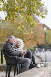 Älteres Paar, das ein digitales Tablet benutzt und Kaffee und Tee im Herbstpark genießt - CAIF26453