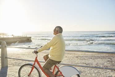 Aktiver älterer Mann, der auf der sonnigen Strandpromenade am Meer Fahrrad fährt - CAIF26494