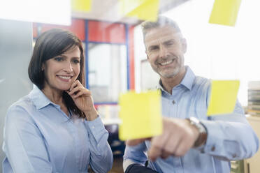 Lächelnde Geschäftsfrau und Geschäftsmann mit Blick auf Haftnotizen in einem Fabrikbüro - DIGF10225