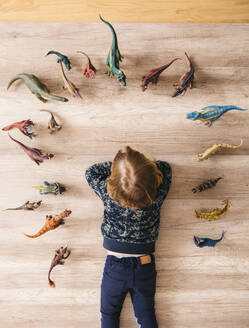 Little girl lying on the floor playing with toy dinosaurs around her, top view - JRFF04407
