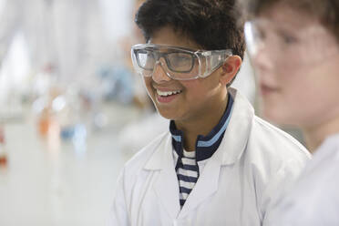 Lächelnder Junge mit Schutzbrille und Laborkittel in einem Laborklassenzimmer - CAIF26520