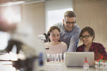 Männlicher Lehrer und weibliche Schüler, die ein wissenschaftliches Experiment an einem Mikroskop und einem Laptop in einem Laborklassenzimmer durchführen - CAIF26530